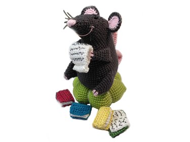 Gehäkelte dunkelgraue Maus mit rosa Ohren auf grünem Kissen, hält weißes gehäkeltes Blatt; bunte Mini‑Bücher davor