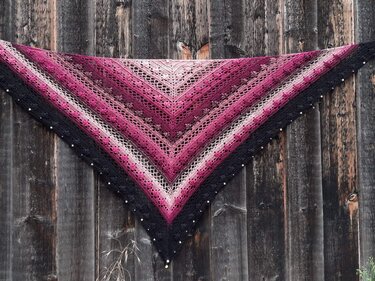 Triangle crochet shawl in pink-to-black gradient with lace and shell stitches and a beaded black border pinned flat on a rustic wooden wall