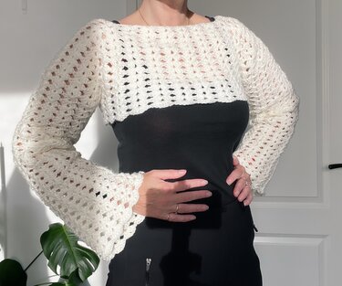 Cropped cream openwork crochet shrug with long flared sleeves, worn over a black top.