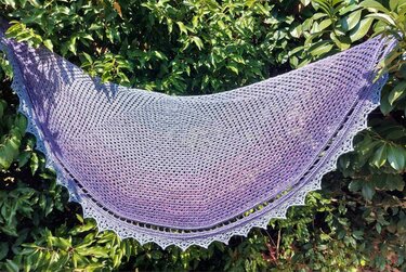 Halbrundes, gehäkeltes Tuch mit Lila‑Blauem Farbverlauf und gezackter Borte, vor grünen Blättern aufgehängt.