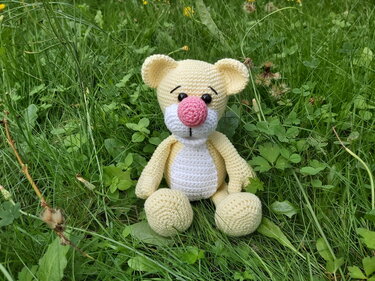 Hellgelber gehäkelter Amigurumi-Teddybär mit weißem Bauch und rosa Nase sitzt im Gras