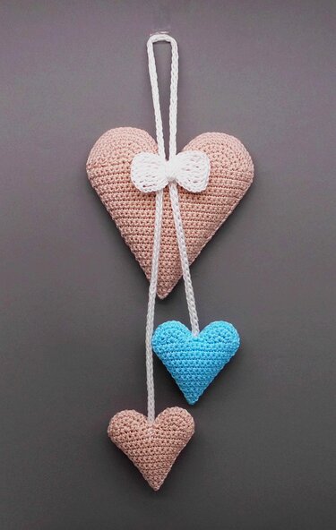 Crocheted hanging decoration with a large pink heart, white bow, and two dangling hearts in blue and pink on a grey background