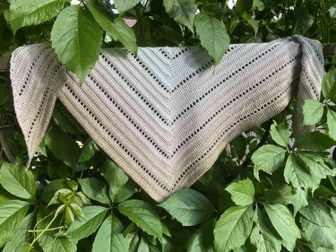 Triangle crochet shawl in neutral variegated yarn with eyelet stripe detail, draped over green leaves