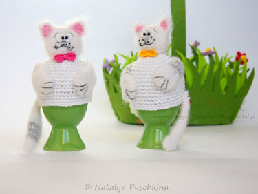 Zwei weiße gehäkelte Katzen-Eierwärmer mit rosa und gelber Schleife auf grünen Eierbechern