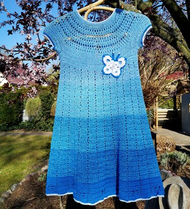 Blaues gehäkeltes Kinderkleid mit weißer Schmetterlings-Applikation an Kleiderbügel vor Garten