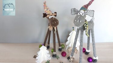 Zwei gehäkelte Elche mit Wichtelmützen und Schals, umgeben von Weihnachtsdekoration auf einem Holztisch.