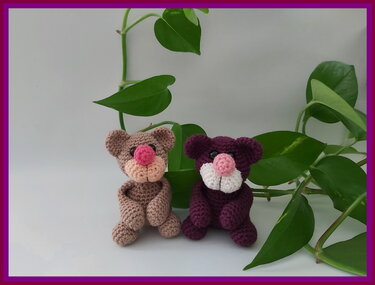 Zwei handgehäkelte Amigurumi-Teddys (braun und lila) mit rosa Nasen vor grünen Blättern