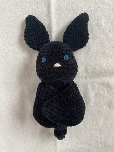 Close-up of a black crocheted bat plush toy with blue eyes and a small pink nose on a light background.