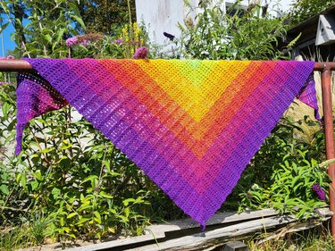 Gehäkeltes Dreieckstuch in Regenbogenfarben, über einer Stange im Garten aufgehängt