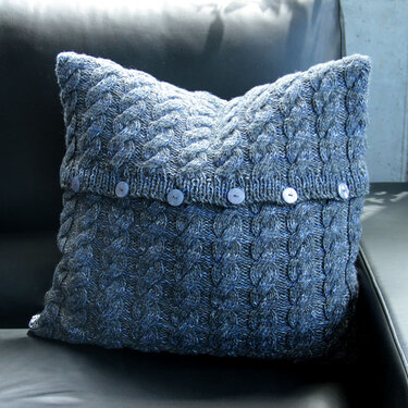 Gestricktes Zierkissen in meliertem Grau‑Blau mit Zopfmuster und Knopfleiste auf dunklem Sofa