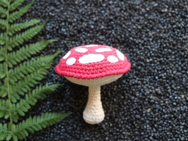 Gehäkelter roter Pilz mit weißen Punkten auf schwarzem Kies neben einem Farnblatt
