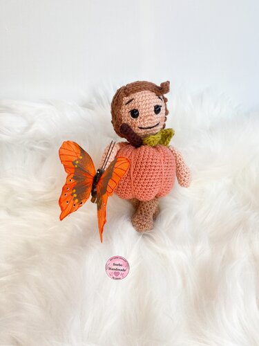Handgehäkelte Amigurumi-Kürbispuppe in Kürbisform mit orangefarbenem Schmetterling auf weißem Fell.