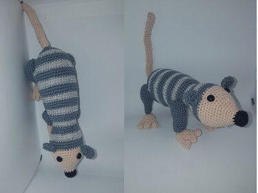 Gehäkeltes Amigurumi-Tier mit gestreiftem Körper in Dunkelgrau und Hellgrau, beige Kopf, Beinen und langem Schwanz.