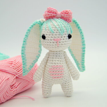 Weißer Amigurumi-Hase mit mintgrünen Ohren, rosa Schleife und rosa Bruststreifen; Garnknäuel im Hintergrund.