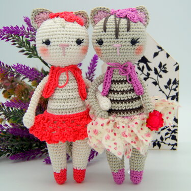 Zwei gehäkelte Amigurumi-Katzenpuppen nebeneinander, eine mit rotem Rock und Halstuch, die andere gestreift mit geblümtem Rock und violettem Halstuch