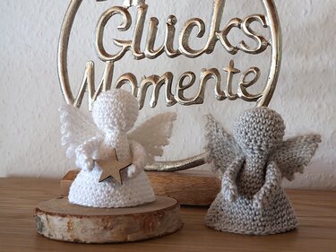 Zwei gehäkelte Amigurumi-Engel, weiß mit Holzstern auf Holzscheibe und grau, vor einem silbernen 'Glücks Momente'-Schild.