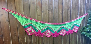 Halbrundes gehäkeltes Tuch in Grün und Pink mit quadratischen Einsätzen und Fransen, aufgehängt an einem Holzzaun