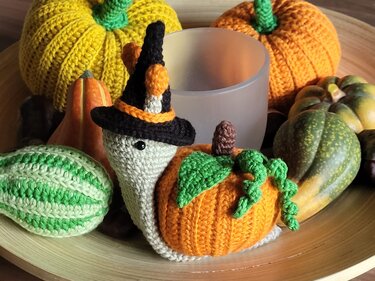 Gehäkelte weiße Schnecke mit orangefarbenem Kürbispanzer, grünen Blattapplikationen und kleinem schwarzem Hexenhut auf einer Holzschale mit Deko-Kürbissen und Kerzenglas