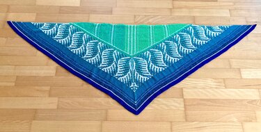 Gestricktes Dreieckstuch blau‑grün mit weißem Blattmuster auf Holzboden