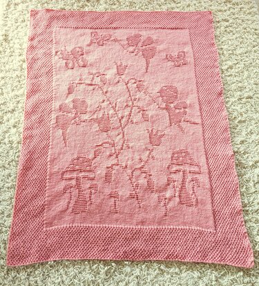 Light pink knitted baby blanket with raised mushroom, butterfly, and floral motifs on a light carpet.