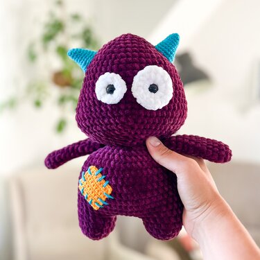 Hand hält lila gehäkeltes Amigurumi-Monster mit weißen Augen, türkisfarbenen Hörnern und orangefarbenem Flicken.