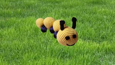 Gehäkelte gelbe Amigurumi-Raupe mit schwarzen Fühlern und lila Beinchen auf grünem Gras