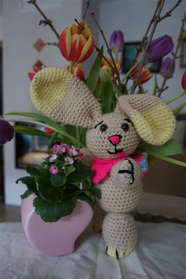 Gehäkeltes beiges Amigurumi-Häschen mit großen Ohren und pinkem Halstuch neben einem pinken Blumentopf und Tulpen