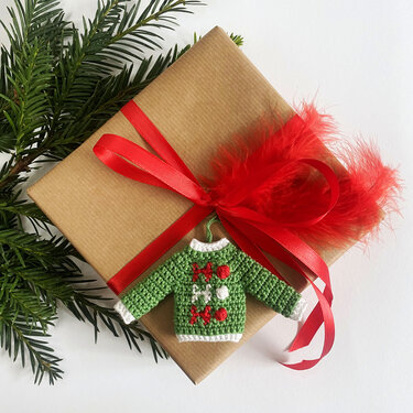 Grüner gehäkelter Mini-Pullover mit 'HO HO HO'-Stickerei als Geschenkanhänger auf braunem Geschenk mit roter Schleife und Tannengrün.