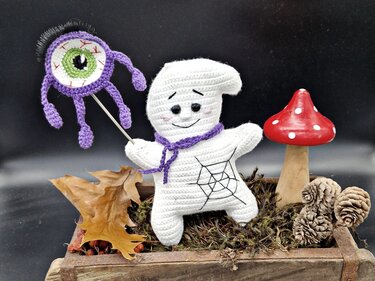 Handgehäkelter weißer Geist mit lila Schal hält lilafarbenes Monsterauge auf Stab, dekoriert mit Moos, Blättern, Tannenzapfen und Fliegenpilz.