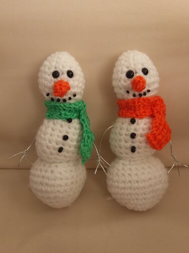 Zwei gehäkelte Amigurumi-Schneemänner mit grünem und orangefarbenem Schal, Perlenaugen und Drahtarmen