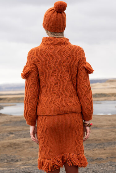 Rückenansicht: Frau in rostorangefarbenem Strickpullover, Rock mit Fransen und Bommelmütze vor einer Naturlandschaft.
