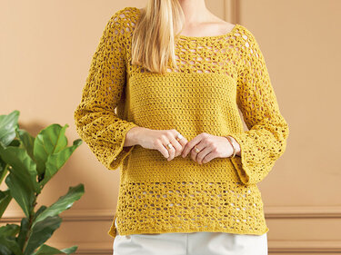 Frau in senfgelbem gehäkeltem Pullover mit Lochmuster und hellen Hosen vor beiger Wand