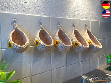 Five crocheted hanging baskets with sunflower appliqués hung on a tiled kitchen wall