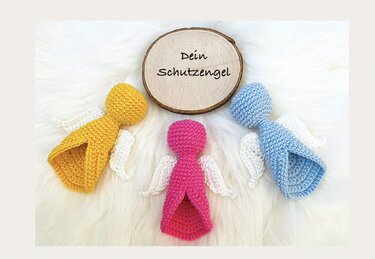 Drei kleine gehäkelte Schutzengel in Gelb, Pink und Hellblau mit weißen Flügeln auf flauschigem Untergrund, mit Holzscheibe „Dein Schutzengel“ in der Mitte.