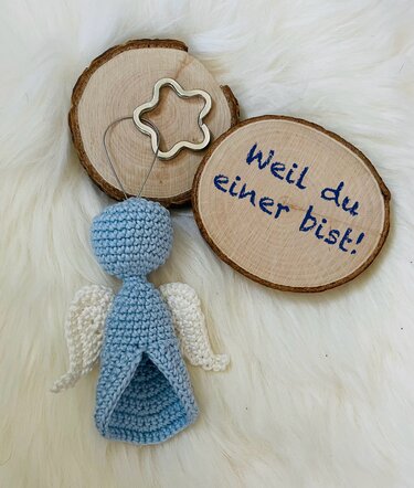 Pastellblaues gehäkeltes Engelchen mit weißen Flügeln auf Fell, Holzscheiben mit handgeschriebener Botschaft