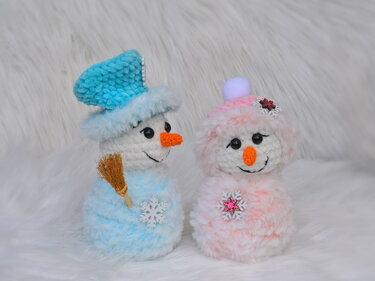 Zwei gehäkelte Amigurumi-Schneemänner in Blau und Rosa auf weißem Plüsch