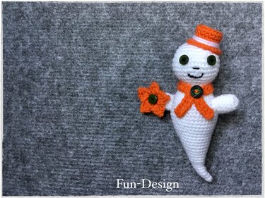 Weißes gehäkeltes Gespenst-Amigurumi mit oranger Mütze, Schal und Blume auf grauem Hintergrund