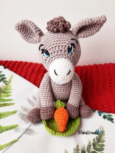 Gehäkelter Amigurumi-Esel sitzt und hält eine Karotte auf grünem Blatt