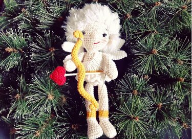 Gehäkelter weißer Amigurumi‑Engel mit gelbem Bogen und rotem Herzpfeil vor Tannenzweigen