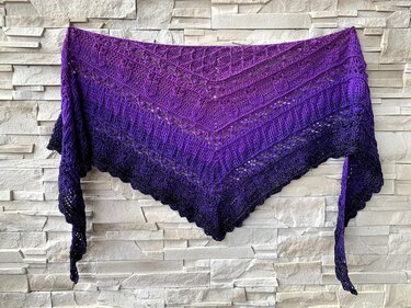 Purple ombre crocheted triangle shawl with lace pattern hanging on a light brick wall
