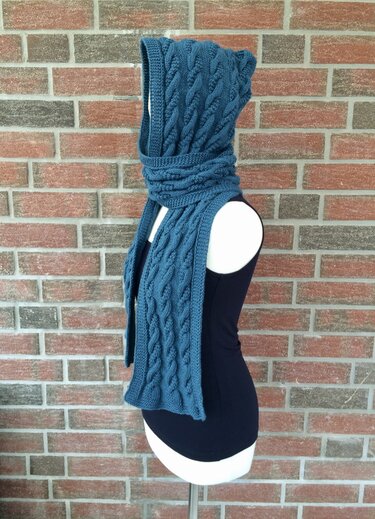 Schaufensterpuppe trägt dunkelblauen gestrickten Kapuzen‑Schal im Zopfmuster vor einer Ziegelwand