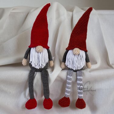 Zwei gehäkelte Wichtel mit roten Zipfelmützen, weißen Bärten und grauen Körpern auf hellem Stoff.
