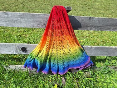 Gestricktes Lacetuch in Regenbogenfarben mit Ajour-Lochmuster, über einem Holzzaun drapiert, Gras im Hintergrund