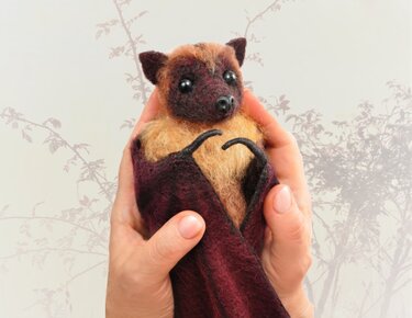 Kleines handgemachtes Fledermaus‑Amigurumi in Braun‑ und Beigetönen, von zwei Händen gehalten vor hellem Hintergrund mit dezenten Pflanzenmotiven.