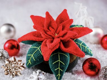 Close-up of a red crocheted poinsettia with embroidered leaf veins and surrounding Christmas baubles