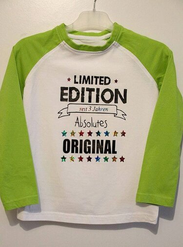 Weißes Kinder-Langarmshirt mit limettengrünen Raglanärmeln und schwarzem Aufdruck "LIMITED EDITION seit 3 Jahren Absolutes ORIGINAL" sowie bunten Sternen, aufgehängt.