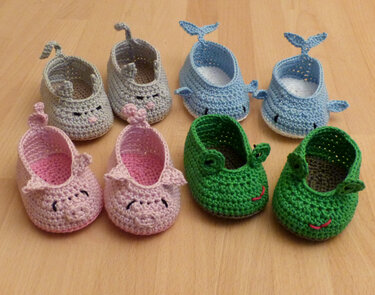 Vier Paar einfache gehäkelte Babyschuhe: blaue Wale, grüne Frösche, rosa Schweinchen und ein graues Paar mit Öhrchen auf hellem Holz