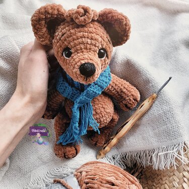 Brauner gehäkelter Amigurumi-Teddy aus Plüschgarn mit blauem Schal, in der Hand gehalten, Häkelnadel und Garn daneben