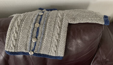 Graue handgestrickte Kinderstrickjacke mit blauen Kanten und metallischen Knöpfen mit floralem Relief, liegend auf einem dunkelbraunen Ledersofa.