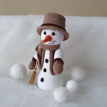 Handgehäkelter Amigurumi-Schneemann mit braunem Zylinder, Schal und Besen, umgeben von weißen Dekoschneebällen.